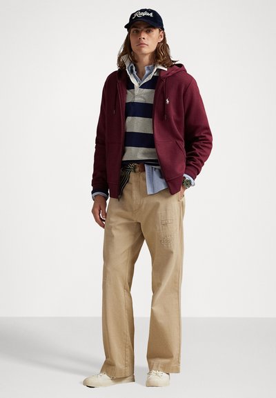 Hoodie zippé bordeaux, chemise rayée grise et marine, sous-vêtement bleu clair, pantalon large en khaki avec des détails texturés, baskets blanches, casquette marine.