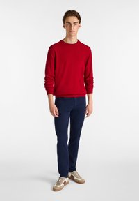 Red sweater with ribbed cuffs and neckline, navy blue slim-fit pants, and beige sneakers with brown accents, standing against a plain background.