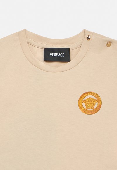 Béžové tričko s kruhovým výstrihom, čiernou štítkou Versace, zlatým emblémom Medusa na hrudi a dvomi zlatými detailmi na gombíky na švových ramenách.