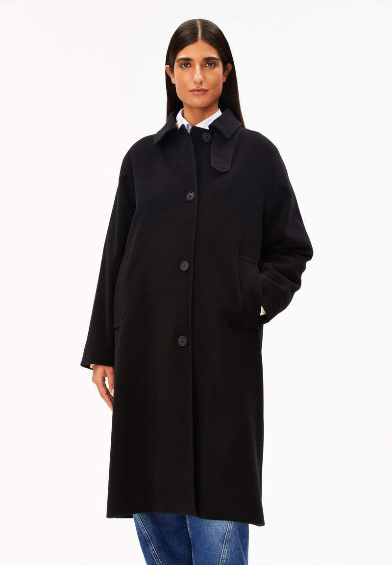 Frau mit einem langen schwarzen knopfbaren Mantel mit Taschen, weißem Kragenhemd und blauen Jeans vor einem schlichten weißen Hintergrund.