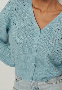 Light blue knitted cardigan with V-neck, button closure, and delicate hole patterns worn over light wash denim jeans.
