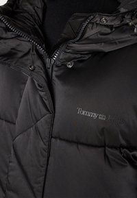 Giubbotto imbottito nero con finitura lucida, caratterizzato da cuciture a forma di diamante, chiusura frontale con zip e un logo sul lato sinistro.