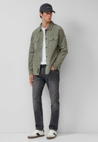 Olivgrüne Cord-Jacke mit zwei Brusttaschen, darunter ein weißes T-Shirt, kombiniert mit dunklen Jeans und weißen Sneakers mit Akzenten.