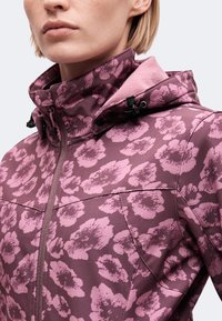 Gros plan d’une personne portant une veste à motifs floraux violets et roses avec un col haut zippé et une capuche ajustable.
