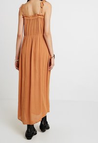 Kvinna som bär en ärmlös, rostfärgad maxiklänning med knytband och svarta ankelstövlar, stående mot en helt vit bakgrund.