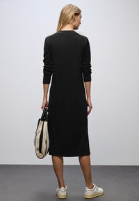 Schwarzes Kleid mit langen Ärmeln und geradem Schnitt, aus weichem Stoff gefertigt. Das Model hält eine beige Tasche mit schwarzen Streifen und trägt weiße Sneakers.
