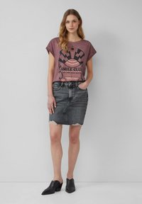 T-shirt graphique en mauve muted avec imprimé noir et manches roulées, associé à une jupe en denim noire avec un ourlet effiloché. Des mules noires complètent le look.