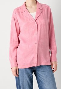 Femme portant une chemise ample rose à boutons avec des motifs subtils de feuilles sur un haut blanc et un jean bleu sur un fond uni.
