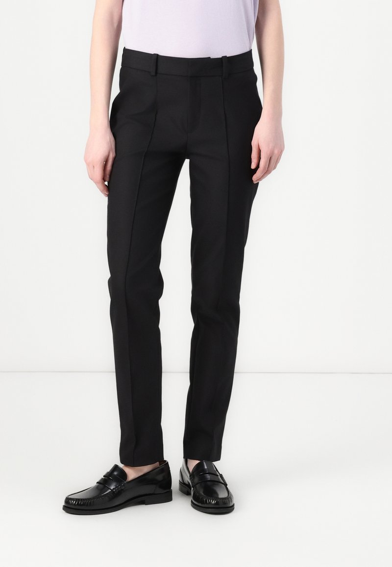Pantalons noirs sur mesure à coupe slim, avec des plis marqués et des passants pour ceinture. Associés à des mocassins noirs brillants.