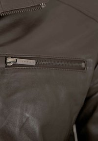 Braune Lederjacke mit glatter Textur, ausgestattet mit einer Reißverschluss-Tasche und einem dezenten Markenetikett in der Nähe des Reißverschlusses.