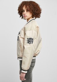 Heledbeez bomber-jope mustade aktsentide ja tikitud logoga varrukal. Omab reljeefseid mansette ja äärt. Tekstureeritud satiinviimistlus.