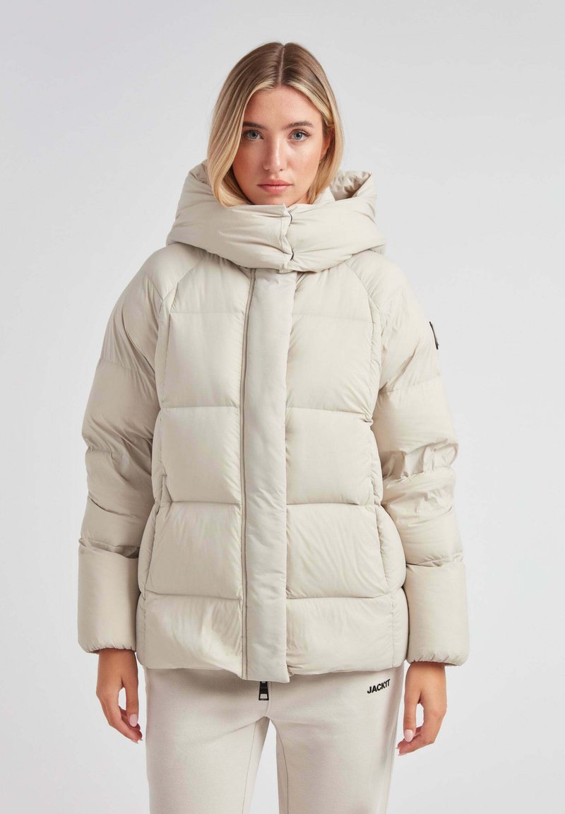 Lys beige pufferjakke med en høj, polstret hætte, quiltet design og lynlås foran. Har et diskret logo nederst på kanten.