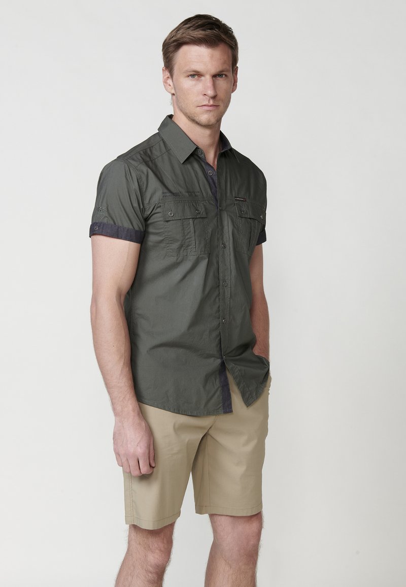 Camisa de botones de manga corta, color verde oliva, con dos bolsillos frontales, acentos oscuros en los puños y una tela ligera, combinada con pantalones cortos beiges.