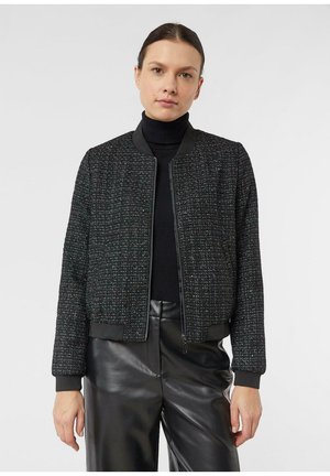 Kobieta ubrana w czarną, teksturowaną kurtkę na zamek, nałożoną na czarny golf, w połączeniu z czarnymi błyszczącymi skórzanymi spodniami, stojąca na jednolitym tle.