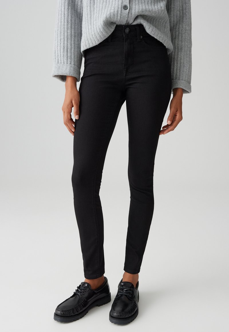 Vrouw die strakke zwarte jeans draagt, een grijze geribbelde trui met opgerolde mouwen en zwarte leren veterschoenen, staand voor een effen achtergrond.