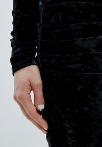 Schwarzes Samtkleid mit strukturierten Oberflächen, das eine figurbetonte Passform und lange Ärmel aufweist, sowie details in den seitlichen Einsätzen.