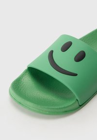 Zöld csúszásmentes szandál matt gumiból. Széles pánttal rendelkezik, amelyen fekete mosolygós arc minta található, valamint texturált talpbetéttel a stabil tapadás érdekében.