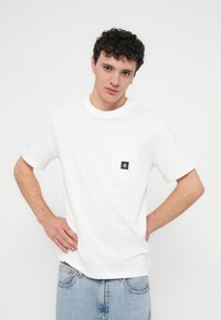 Λευκό βαμβακερό T-shirt με κοντά μανίκια, στρογγυλή λαιμόκοψη και αριστερή τσέπη στο στήθος με μαύρο λογότυπο. Φορεμένο με ανοιχτό μπλε τζιν.