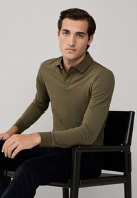 Mladý muž s tmavými vlasmi, oblečený v olivovo zelenom dlhom polo tričku, sedí na čiernej stoličke pred jednoduchým pozadím.