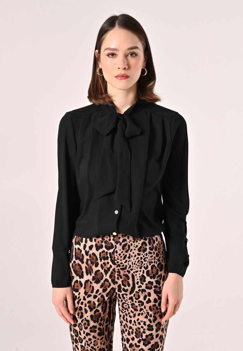 Blusa nera con colletto a fiocco e maniche lunghe, abbinata a pantaloni beige con stampa leopardata. Tessuto morbido, design aderente.