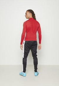 Rotes langärmliges Sportoberteil kombiniert mit schwarzen Hosen, die weiße seitliche Streifen haben. Das Model trägt blaue Sportschuhe mit gelben Akzenten.