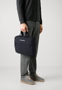 Svart nylon laptopveske med to håndtak og en glidelåslomme foran. Merkenavnet "KARL LAGERFELD" er trykt i hvitt på forsiden.