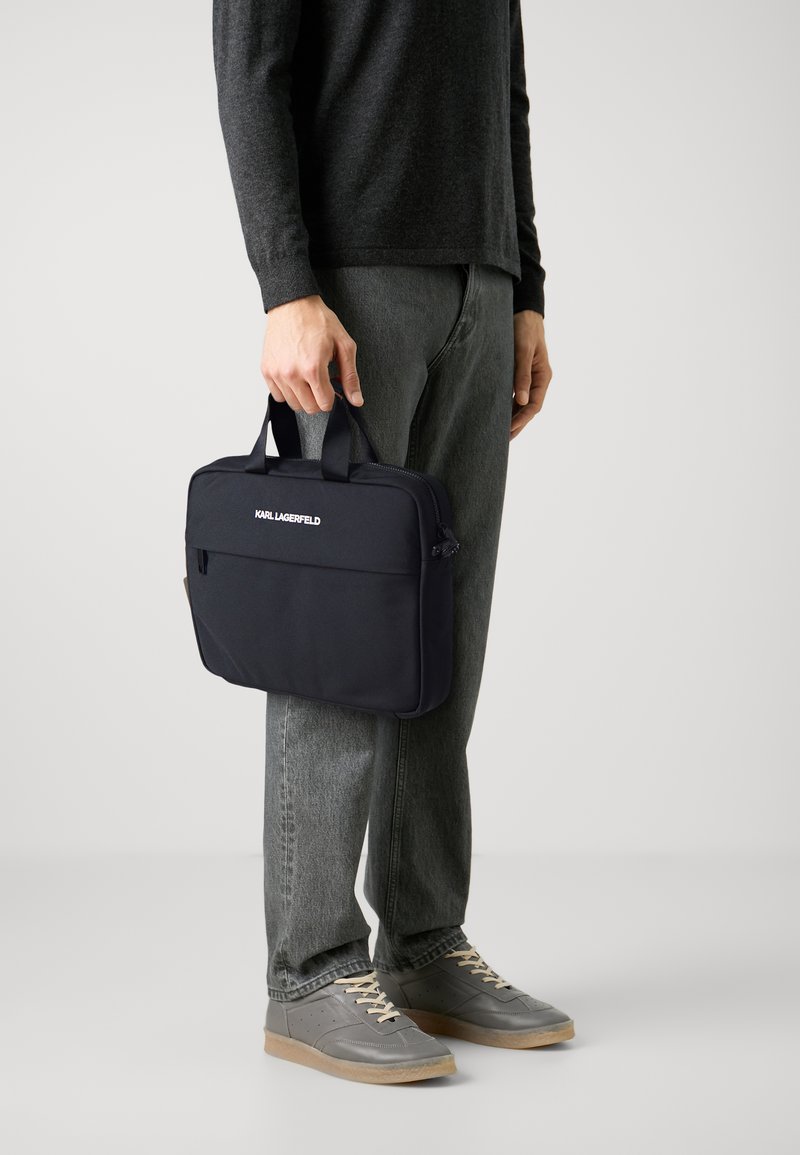 Svart nylon laptopveske med to håndtak og en glidelåslomme foran. Merkenavnet "KARL LAGERFELD" er trykt i hvitt på forsiden.