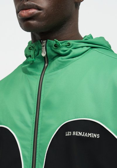 Κοντινό πλάνο ενός ατόμου που φοράει πράσινο και μαύρο μπουφάν με φερμουάρ και κουκούλα, με το μήνυμα «LES BENJAMINS» κεντημένο στο στήθος.