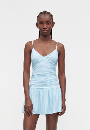 Frau mit geflochtenem Haar, die ein hellblaues, ärmelloses, gerafftes Mini-Kleid mit dünnen Trägern und dezenten vertikalen Streifen trägt und vor einem schlichten Hintergrund steht.