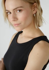 Zwarte geribde tanktop met een brede halslijn en mouwloos ontwerp. Model heeft kort blond haar en zilveren creolen.