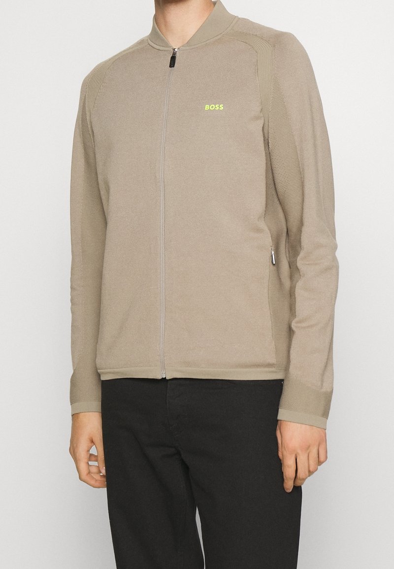 Béžová bunda na zip s vysokým límcem, texturovanou látkou a žebrovanými detaily. Na hrudi malý zelený nápis "BOSS".