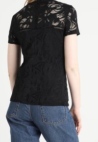 Černý krajkový top s krátkými rukávy, zdobený květinovým vzorem a texturovaným povrchem. Navržený s přiléhavým střihem a zaobleným zadním lemem.