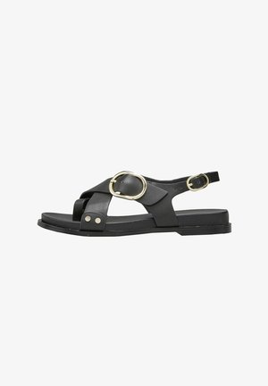 Sandale en cuir noir avec un design à lanières croisées, deux boucles circulaires en or et une semelle plate. Présente des accents de clous en ton argent sur les lanières.