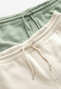 Deux paires de pantalons de jogging : l'une en vert clair et l'autre en crème. Les deux présentent des ceintures élastiques et des cordons de serrage avec des embouts en métal.