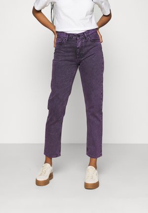 Persona con jeans morados de cintura alta y zapatillas blancas, de pie con las manos detrás de la espalda contra un fondo claro y sencillo.