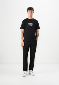 Black cotton t-shirt with graphic print on chest, paired with black trousers and white sneakers. Straight-cut design, minimal texture.