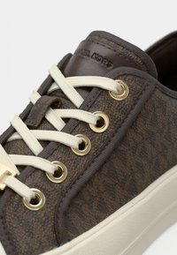 Zapatilla con patrón marrón y material texturizado, cordones crema, ojales dorados y suela de goma blanca contrastante. Logotipo en relieve en la lengüeta.