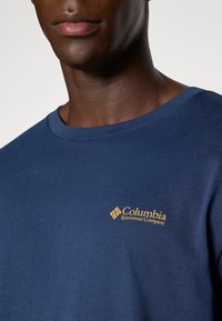 Tumesinine puuvillane T-särk ümarate kaelustega ja lühikeste varrukatega. Ees on kuldne logo Columbia Sportswear Company.