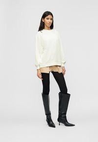 Γυναίκα που φοράει λευκό φαρδύ πουλόβερ, μπεζ σορτς, μαύρο καλσόν και μαύρες μπότες με τακούνι μέχρι το γόνατο, στέκεται μπροστά σε φωτεινό απλό φόντο.