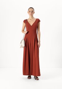 Jumpsuit boje hrđe s dubokim V-izrezom, kratkim rukavima i nabranim dizajnom. Uparen s malom bijelom torbicom i crnim sandalama s trakama.