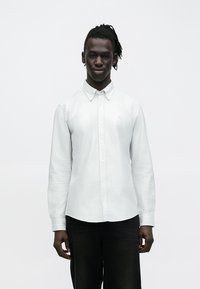 Camisa de manga larga con rayas blancas y cuello abotonado, combinada con jeans oscuros. Presenta una textura sutil y un diseño ajustado.