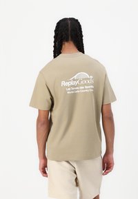 T-shirt in cotone khaki a maniche corte, con testo bianco sul retro: "Replay Goods" e "Monte-Carlo Country Club." Design semplice.