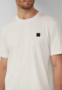 Witte katoenen t-shirt met een ronde hals en korte mouwen, voorzien van een klein zwart rechthoekig logopatch op de linkerborst.