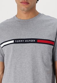 Grå bomulds t-shirt med et horisontalt stribedesign i marineblå, hvid og rød, samt "TOMMY HILFIGER" tekst i midten.