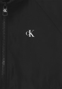 Černá bunda s hladkou texturou, s zipem na přední straně a výrazným bílým logem "cK" na levé straně.