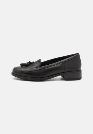Mocasín de piel negra con punta redondeada, detalle decorativo de borlas en la parte delantera y un tacón bajo cuadrado. Textura suave y diseño minimalista.