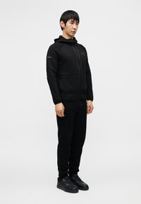 Μαύρο φούτερ με φερμουάρ μπροστά και τσέπη, φορεμένο με μαύρο τζόκερ και μαύρα αθλητικά παπούτσια. Το ύφασμα φαίνεται λείο με ελάχιστες λεπτομέρειες.