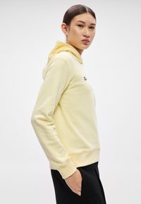 Žltá mikina s kapucňou a mäkkou textúrou, s klokankovým vreckom, rebrovanými manžetami a kapucňou so sťahovacou šnúrkou. Čierny grafický text na prednej strane.