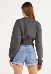 Nainen, jolla on pitkät ruskeat hiukset, pukeutuneena tummanharmaaseen lyhytlahkeiseen huppariin ja vaaleansinisiin käännettyhalkaisijalla denim-shorteihin, kääntyy poispäin kamerasta.