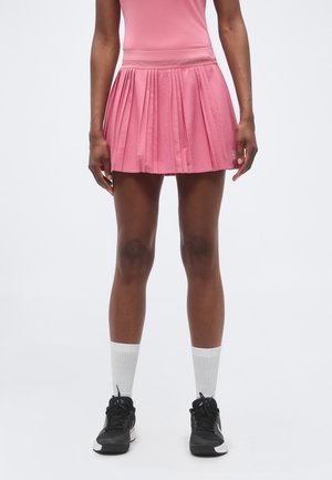 Femeie purtând fustă de tenis plisată roz, șosete albe și pantofi sport negri, stând în fața unui fundal deschis simplu.